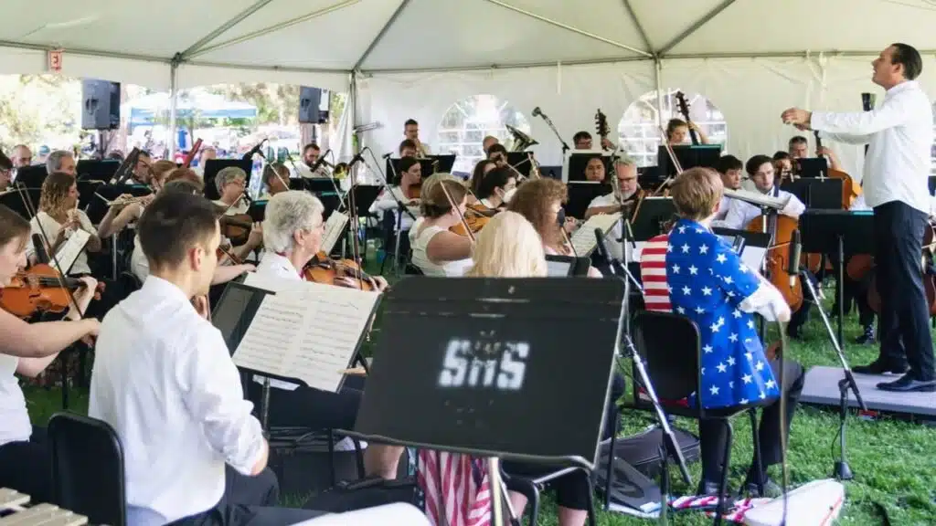 Longmont Fourth of July Concert Returns to Roosevelt Park in 2025 A conductor leads the Longmont Symphony Orchestra during a Fourth of July concert in Roosevelt Park, with musicians performing under a white event tent.