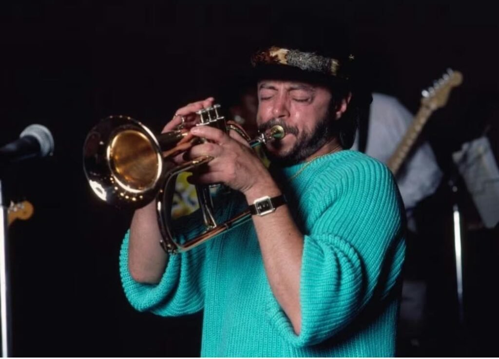 Jazz musician Chuck Mangione performing live on flugelhorn while wearing a teal sweater and patterned cap, captured during a concert in the 1980s.