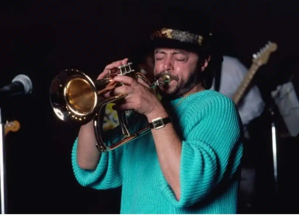 Chuck Mangione Death: Jazz Legend Dies at 84 in Rochester, NY Jazz musician Chuck Mangione performing live on flugelhorn while wearing a teal sweater and patterned cap, captured during a concert in the 1980s.