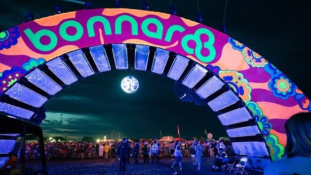 Bonnaroo 2026 Returns to Manchester After 2025 Weather Cancellation Crowds gather under the colorful, psychedelic Bonnaroo arch at night with a disco ball hanging above.
