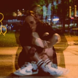 Indie artist mon cher sitting outdoors at night, double exposure effect with blurred shadows, wearing white Nike sneakers.