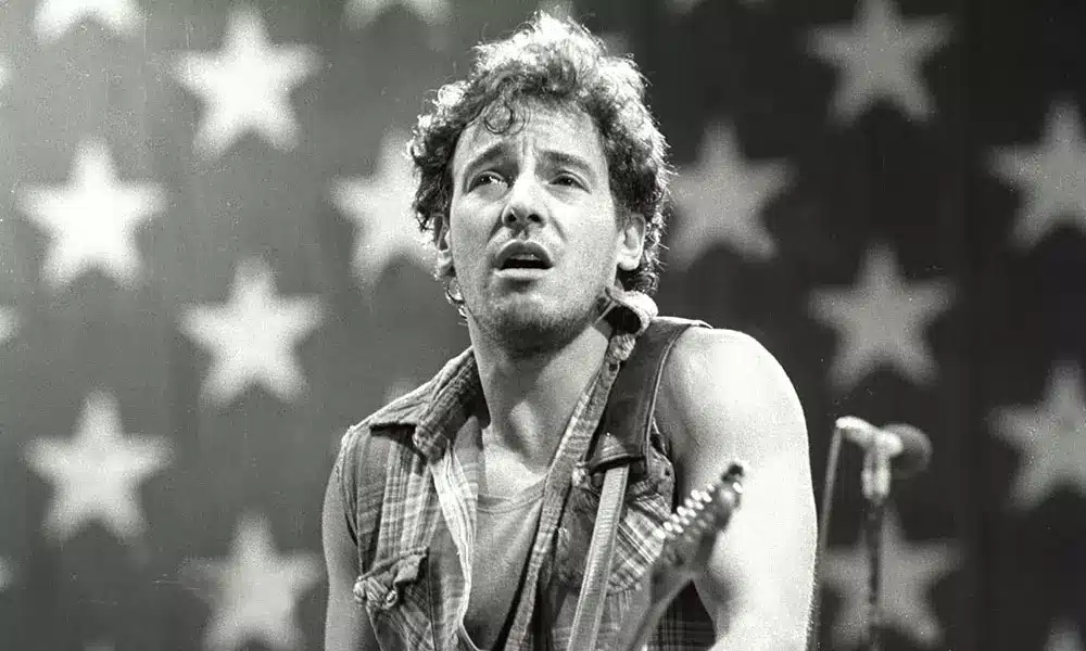 Bruce Springsteen performing live in front of an American flag backdrop, wearing a sleeveless shirt and playing guitar.