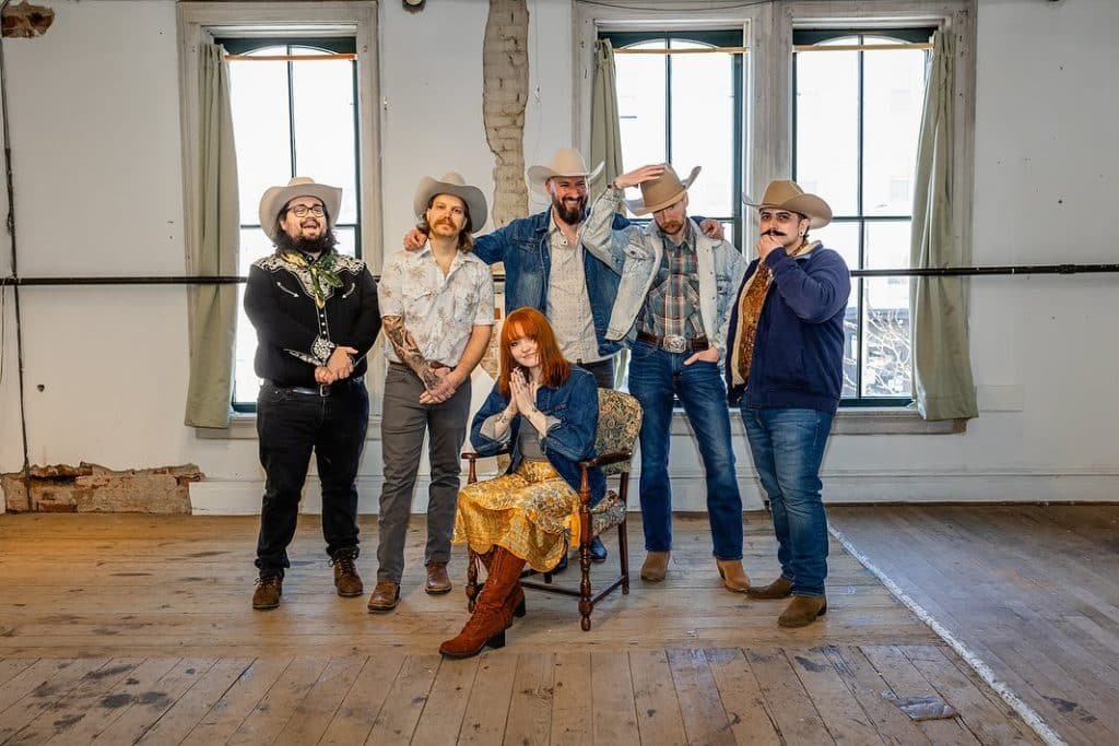 Six members of the Sugar Britches denver band posing in a rustic studio with wooden floors and large windows; five men wearing cowboy hats and western denim attire stand behind a woman seated in a vintage chair wearing a denim jacket.