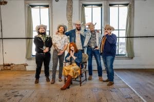 Six members of the Sugar Britches denver band posing in a rustic studio with wooden floors and large windows; five men wearing cowboy hats and western denim attire stand behind a woman seated in a vintage chair wearing a denim jacket.
