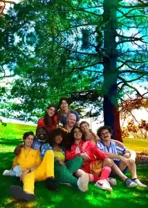 Full 9-piece lineup of the DOGTAGS band sitting together on green grass under a large tree, featuring founders Regi Worles and Michael Merola.