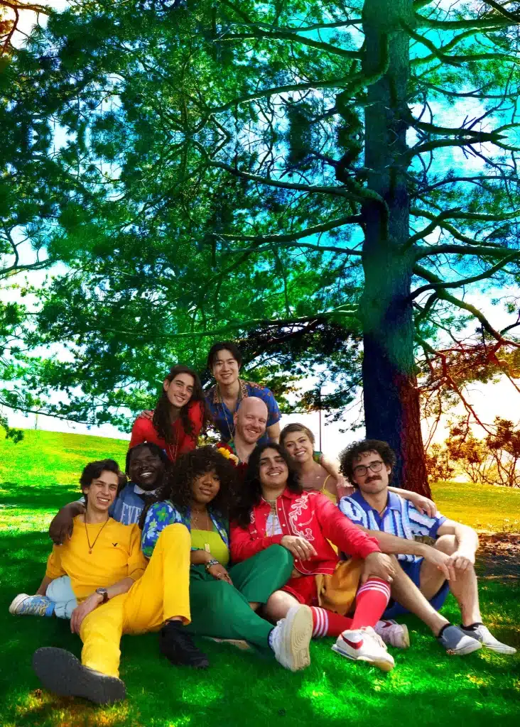 Full 9-piece lineup of the DOGTAGS band sitting together on green grass under a large tree, featuring founders Regi Worles and Michael Merola.