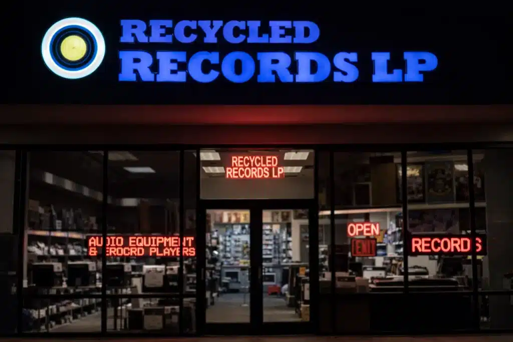 Exterior view of Recycled Records LP, a local record store, at night with illuminated signs and records visible inside.