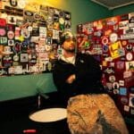 Keenan TreVon, an Aurora-born R&B artist, wearing a black beanie and hoodie, leaning against a sticker-covered wall in a visually textured room.