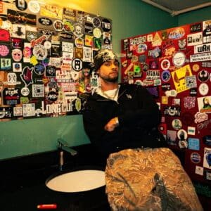 Keenan TreVon, an Aurora-born R&B artist, wearing a black beanie and hoodie, leaning against a sticker-covered wall in a visually textured room.