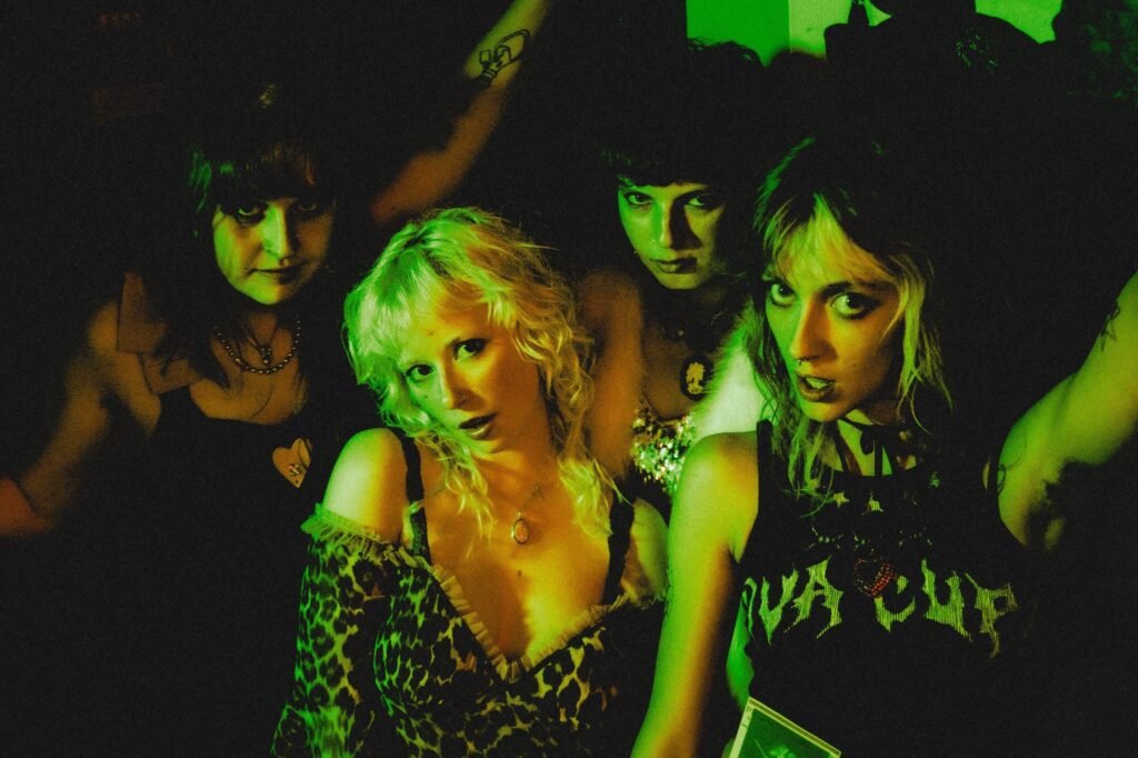 The four members of the Diva Cup band in Denver posing under green stage lights, wearing punk-inspired clothing including a leopard print top and a DIY Diva Cup shirt.