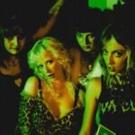 The four members of the Diva Cup band in Denver posing under green stage lights, wearing punk-inspired clothing including a leopard print top and a DIY Diva Cup shirt.